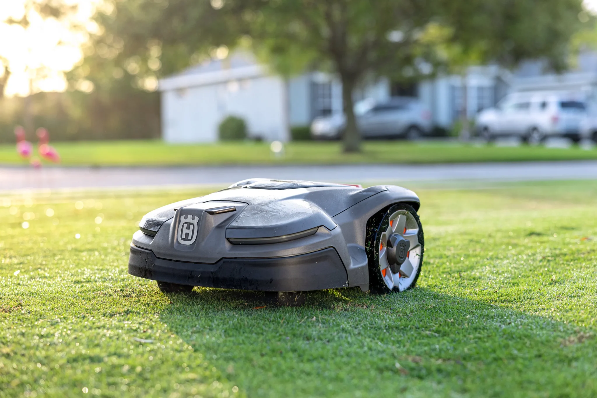 En robotgräsklippare med Husqvarnas logotyp rör sig över en gräsmatta med hus, träd och parkerade bilar suddiga i bakgrunden.
