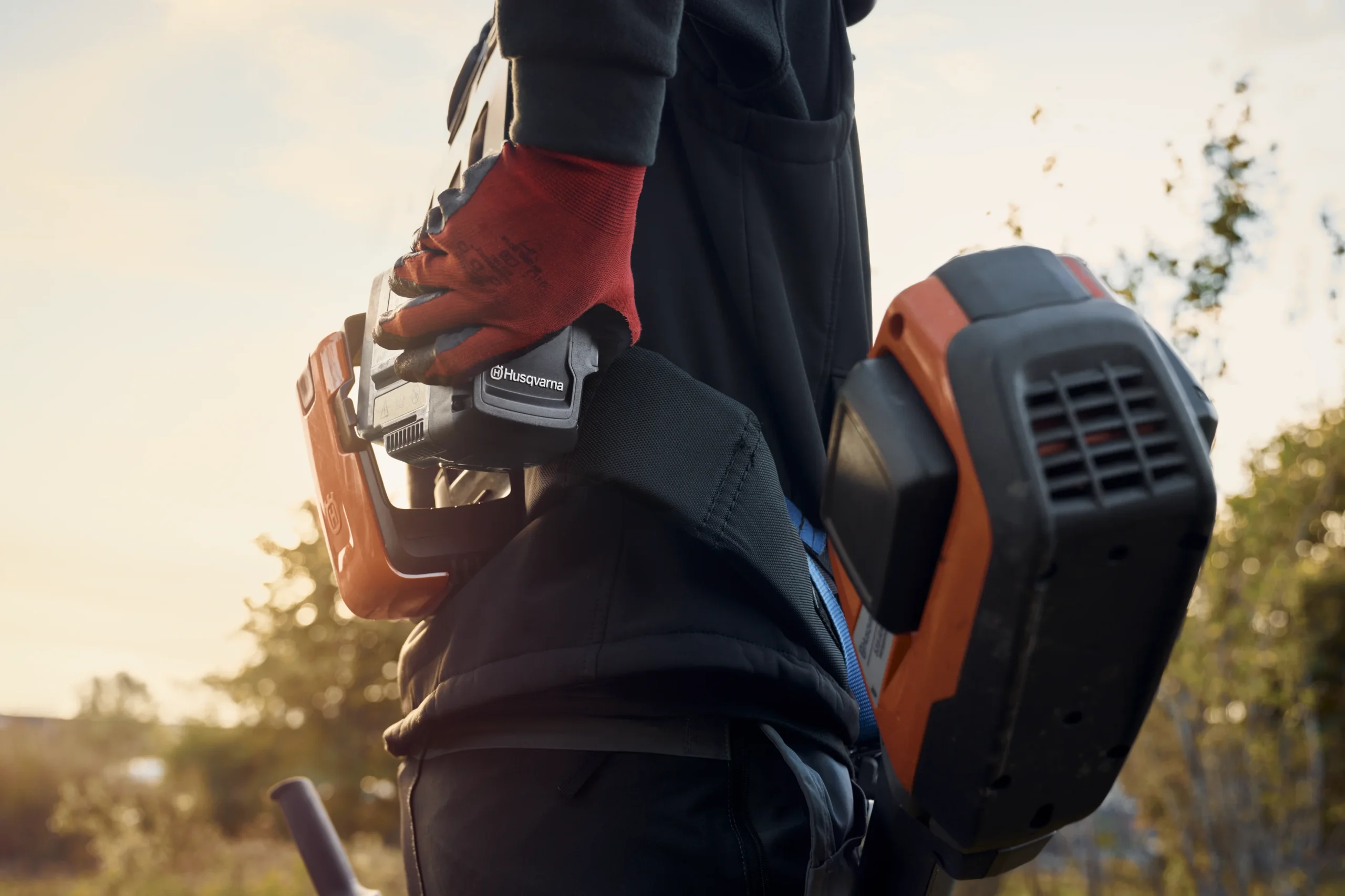 SI-410916 En person med röda handskar håller i ett Husqvarna-batteri, fastspänd med elutrustning och verktyg i midjan, stående utomhus med solljus i bakgrunden.