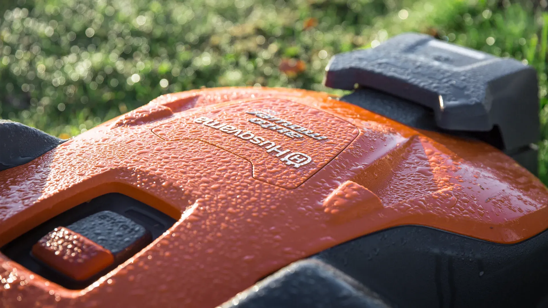 Närbild av en daggtäckt orange Husqvarna robotgräsklippare med gräs suddigt i bakgrunden. Husqvarnas logotyp och modellinformation syns på maskinens yta.