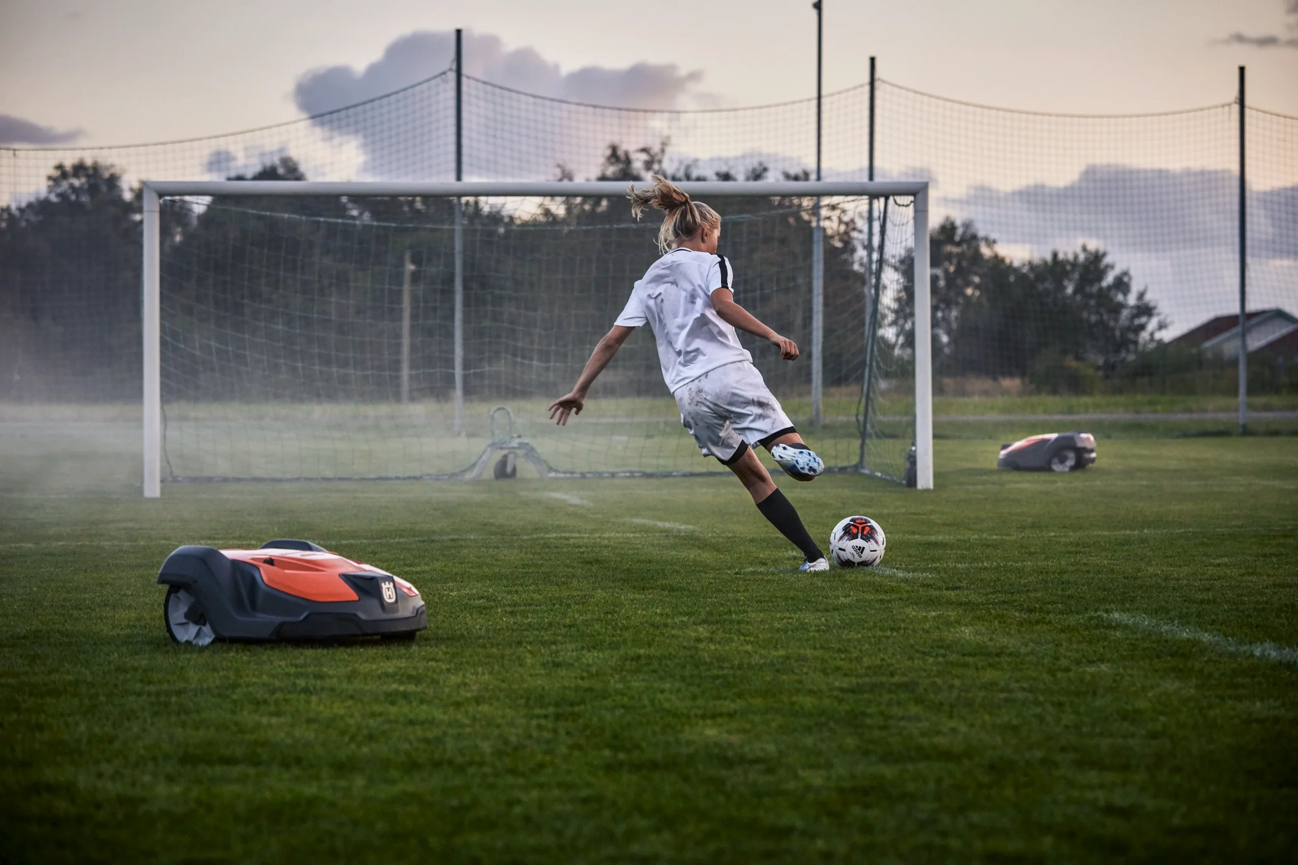 H350-0770 En ung fotbollsspelare i vit uniform sparkar en boll mot ett mål på en gräsplan, medan två robotgräsklippare lugnt sköter om gräset i närheten.