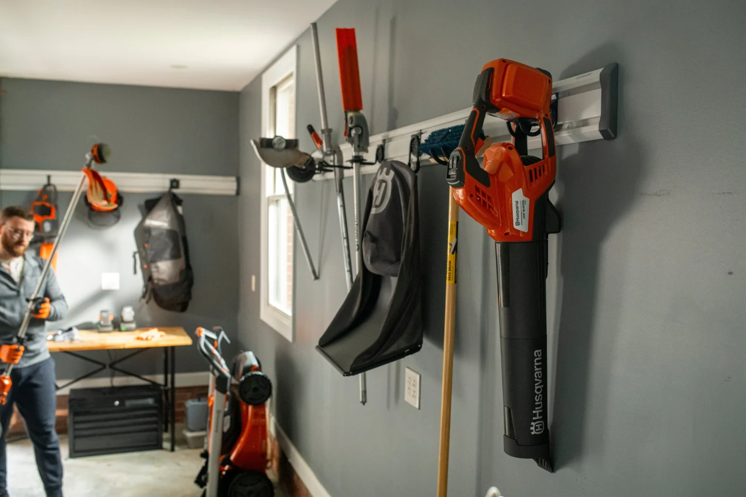 Ett garage med orange och svarta Husqvarna trädgårdsredskap och utrustning, inklusive en robotgräsklippare för idrottsföreningar, organiserade på väggmonterade ställningar. En person håller i en trimmer i bakgrunden nära en arbetsbänk med trädgårdsredskap.