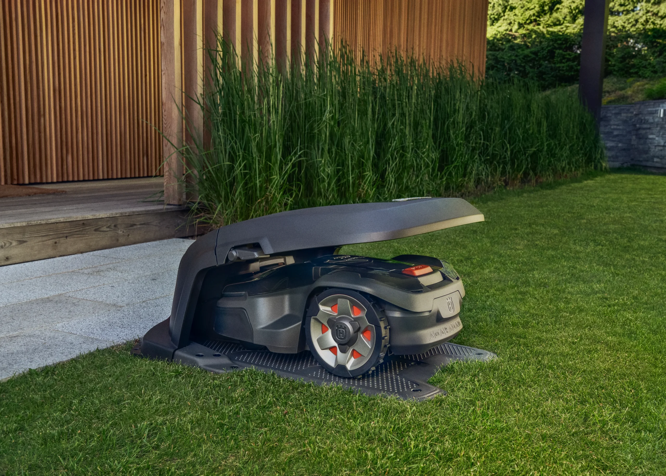En robotgräsklippare sitter på sin laddstation under ett öppet skyddshölje, placerad i utkanten av en välklippt gräsmatta bredvid en modern träbyggnad med höga gröna växter i bakgrunden.