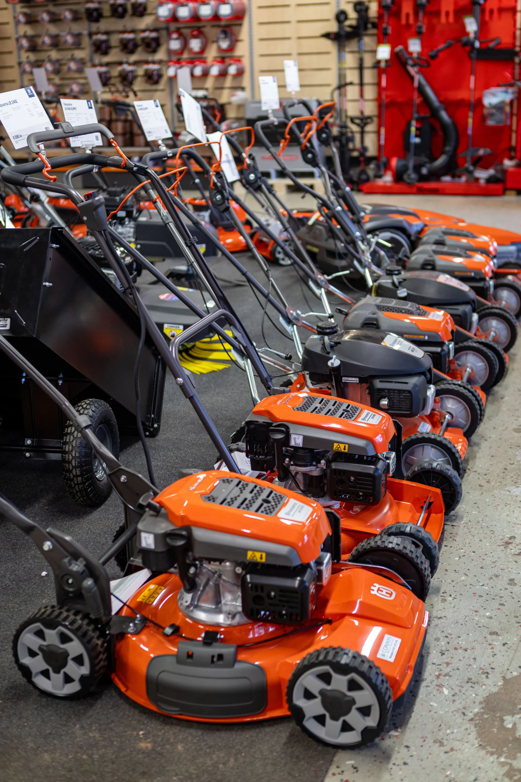 En rad orangefärgade gräsklippare står uppradade i en järnaffär, med olika modeller utställda på ett utställningsgolv bredvid en vägg med trädgårdsredskap - läs mer om vårt urval under "om oss.
